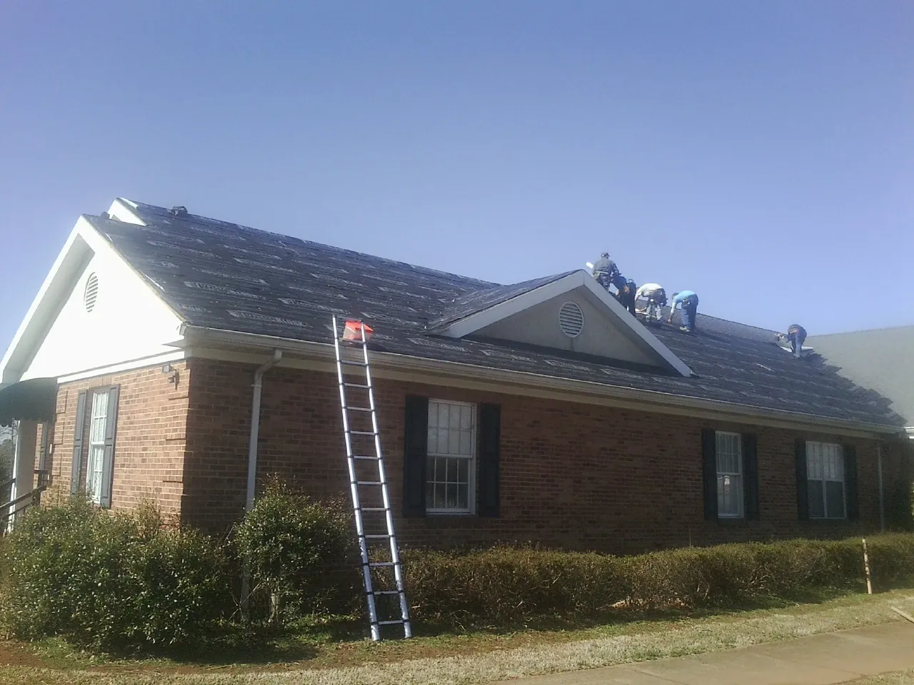 Crew tearing shingles off a brick ranch roof with a ladder set — Metal Roof Repair work by our crew in Philo