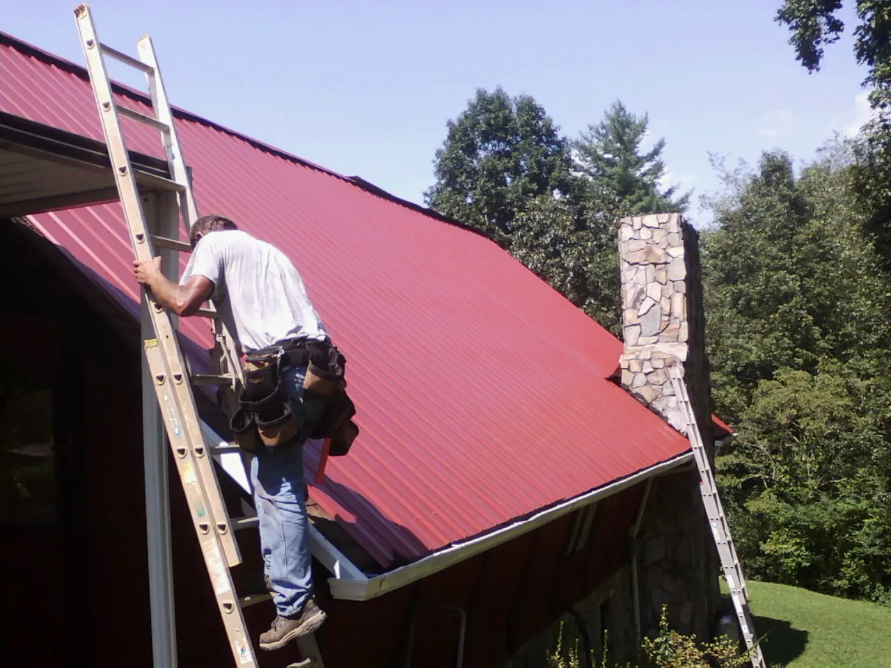 Roofer on a ladder beside a red metal roof and stone chimney — Soffit & Fascia Repair by Roofing Brothers in Philo